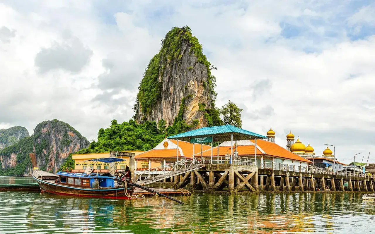 Scogliera verdeggiante su un'isola, con un villaggio su palafitte e un molo. Sembra l'isola di James Bond, con barche tradizionali che navigano sull'acqua.