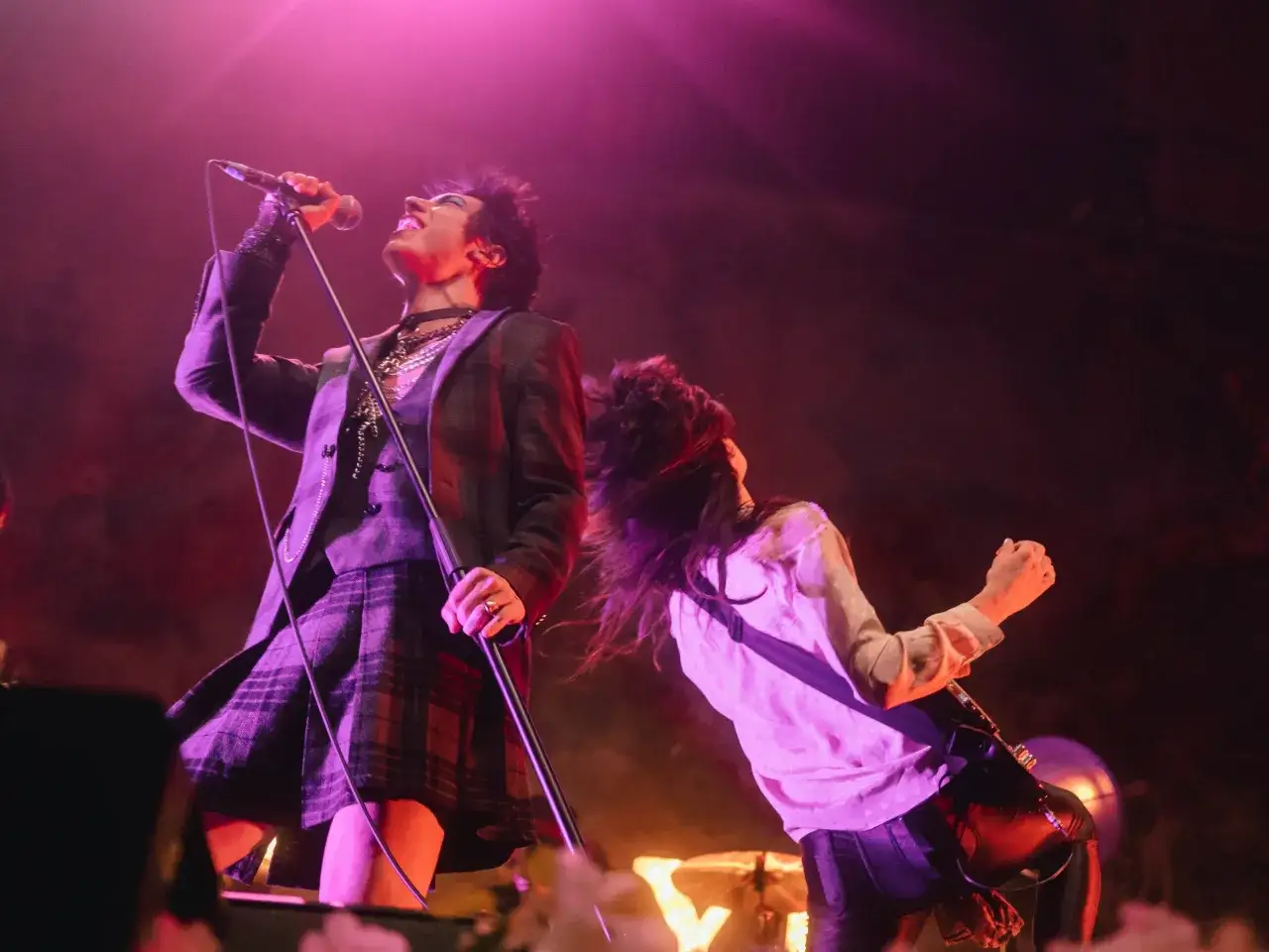 Palaye Royale członkowie na scenie: wokalista w kraciastej spódniczce i marynarce śpiewa do mikrofonu, a gitarzysta z burzą włosów szaleje przy gitarze.