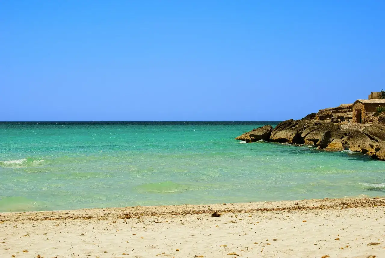T&uuml;rkisfarbenes Meer und Sandstrand in Portocolom, Mallorca. Felsen mit einem kleinen Steingeb&auml;ude ragen ins Wasser.