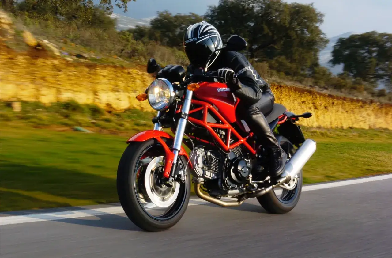 Czerwony Ducati Monster 696 z kierowcą w kasku i skórze pędzi po krętej drodze wśród zieleni i żółtych skarp.