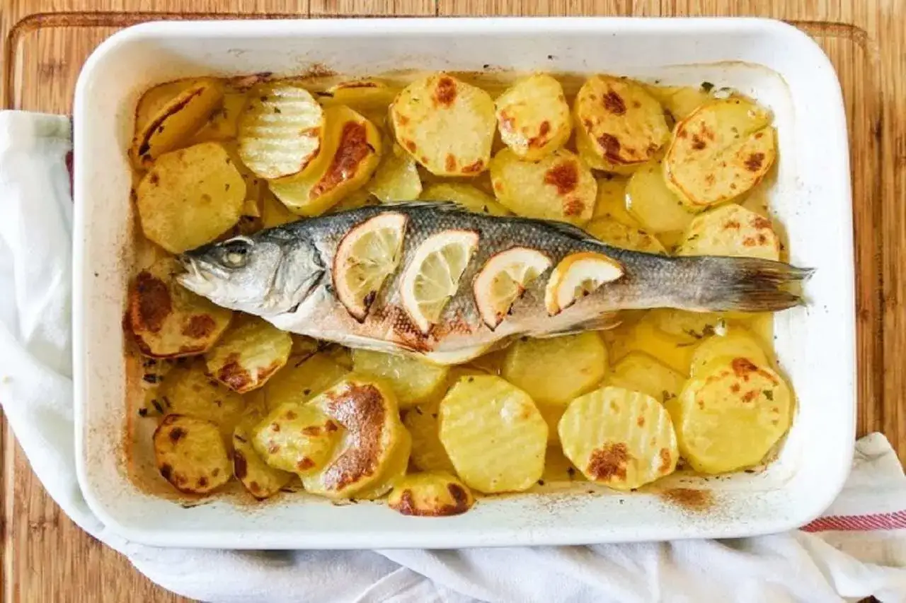 Un intero branzino al forno, adagiato su patate dorate e affettate, guarnito con spicchi di limone.