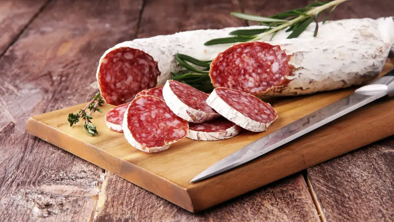 Salami auf einem Holzbrett, daneben ein Messer und Kräuter. Darf man in der Schwangerschaft Salami essen? Diese Frage beschäftigt viele werdende Mütter.