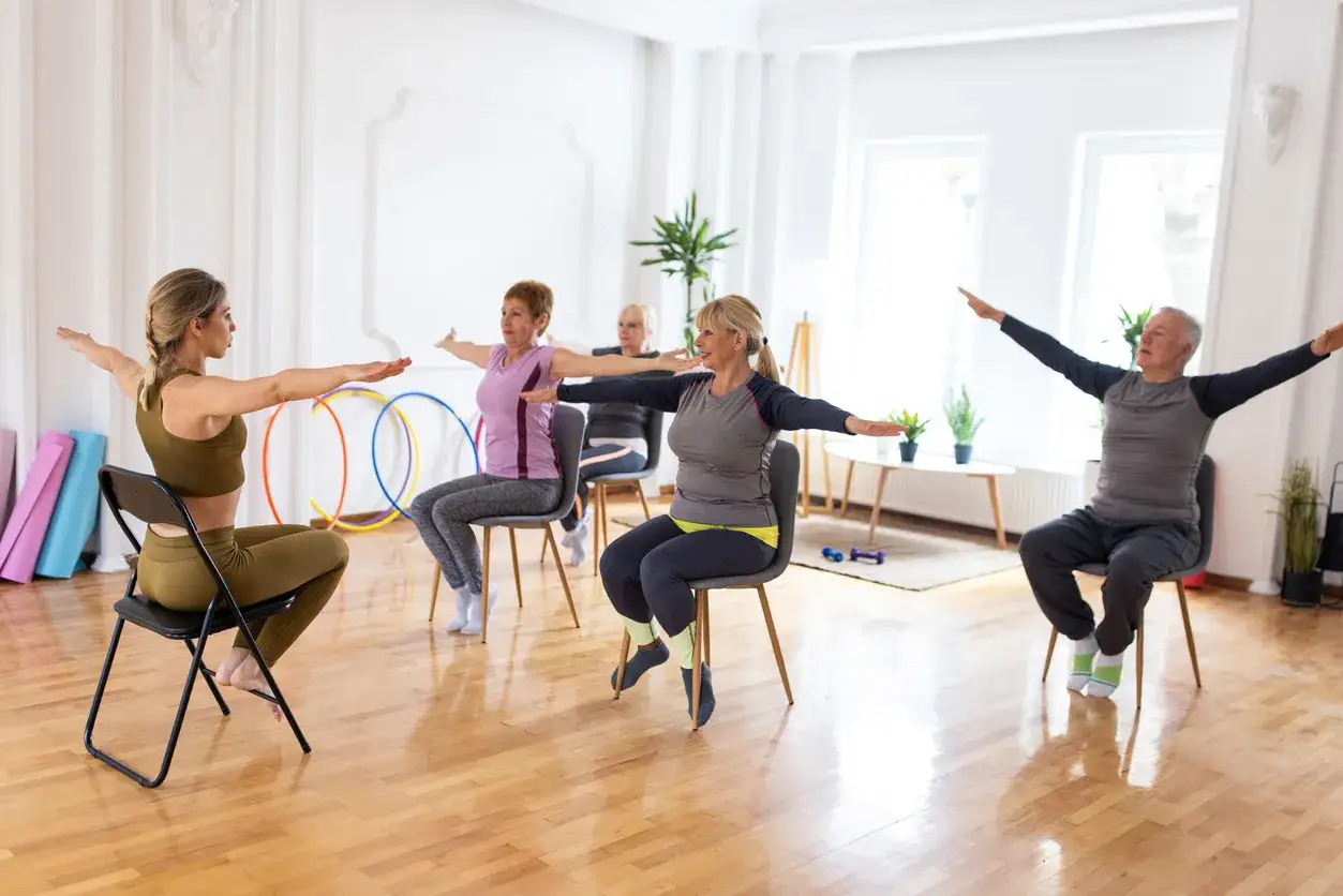 Grupa seniorów wykonuje ćwiczenia na krześle pod okiem instruktorki. Wszyscy mają rozłożone ręce.