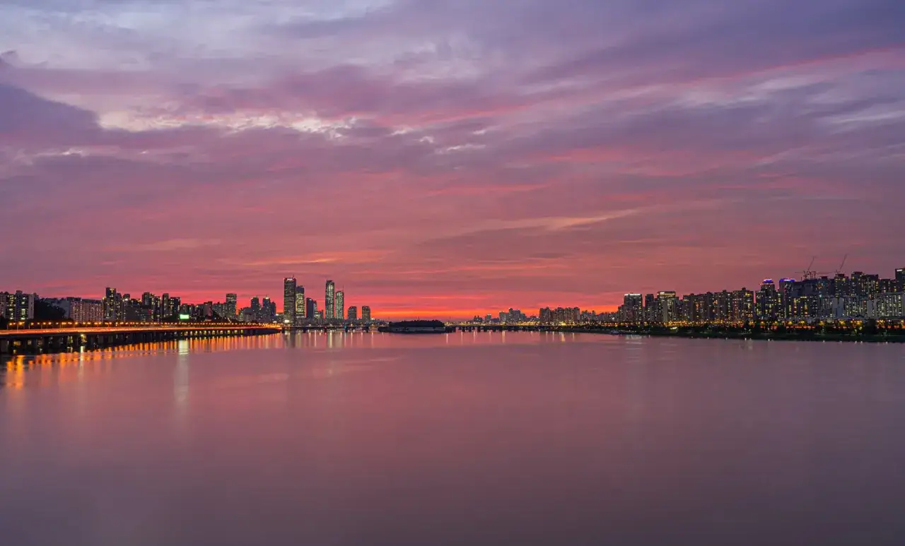 En dramatisk solnedg&aring;ng &ouml;ver floden Han i Seoul, Sydkorea, med stadens silhuett och upplysta broar som speglas i vattnet.