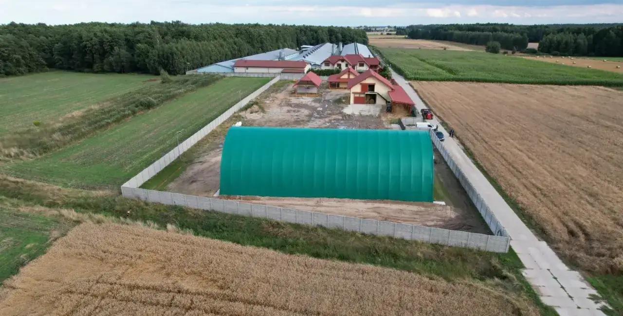 Zielony namiot magazynowy na działce rolnej, obok budynków gospodarczych i pól uprawnych. Widać ogrodzenie i drogę dojazdową.