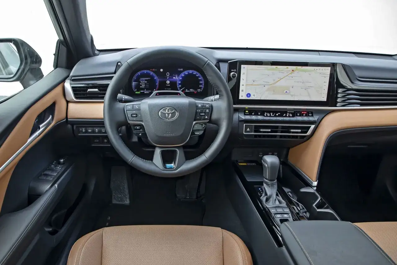 Wnętrze Toyota Camry 2.5 Hybrid: kierownica, ekran nawigacji, cyfrowe zegary i brązowa skórzana tapicerka.
