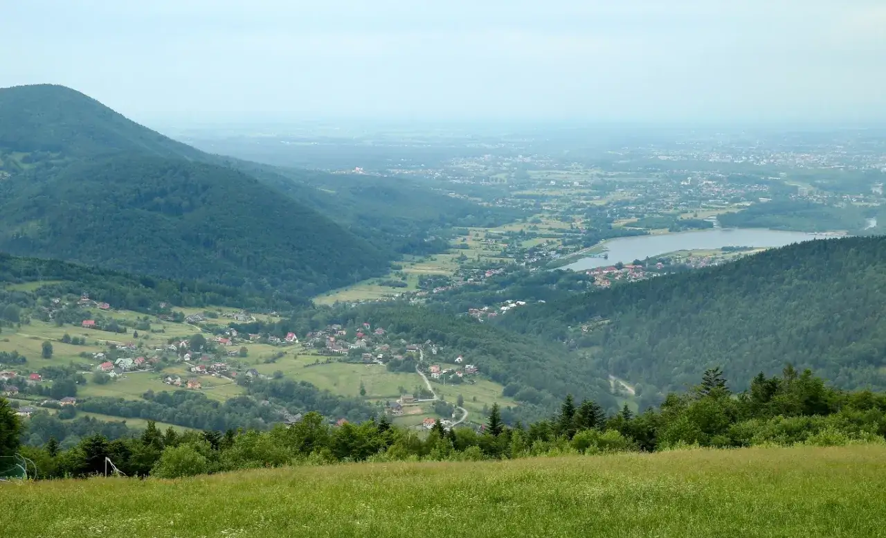 Widok na dolinę z jeziorem i zabudowaniami, otoczoną zielonymi wzgórzami. To piękny fragment Małego Szlaku Beskidzkiego.