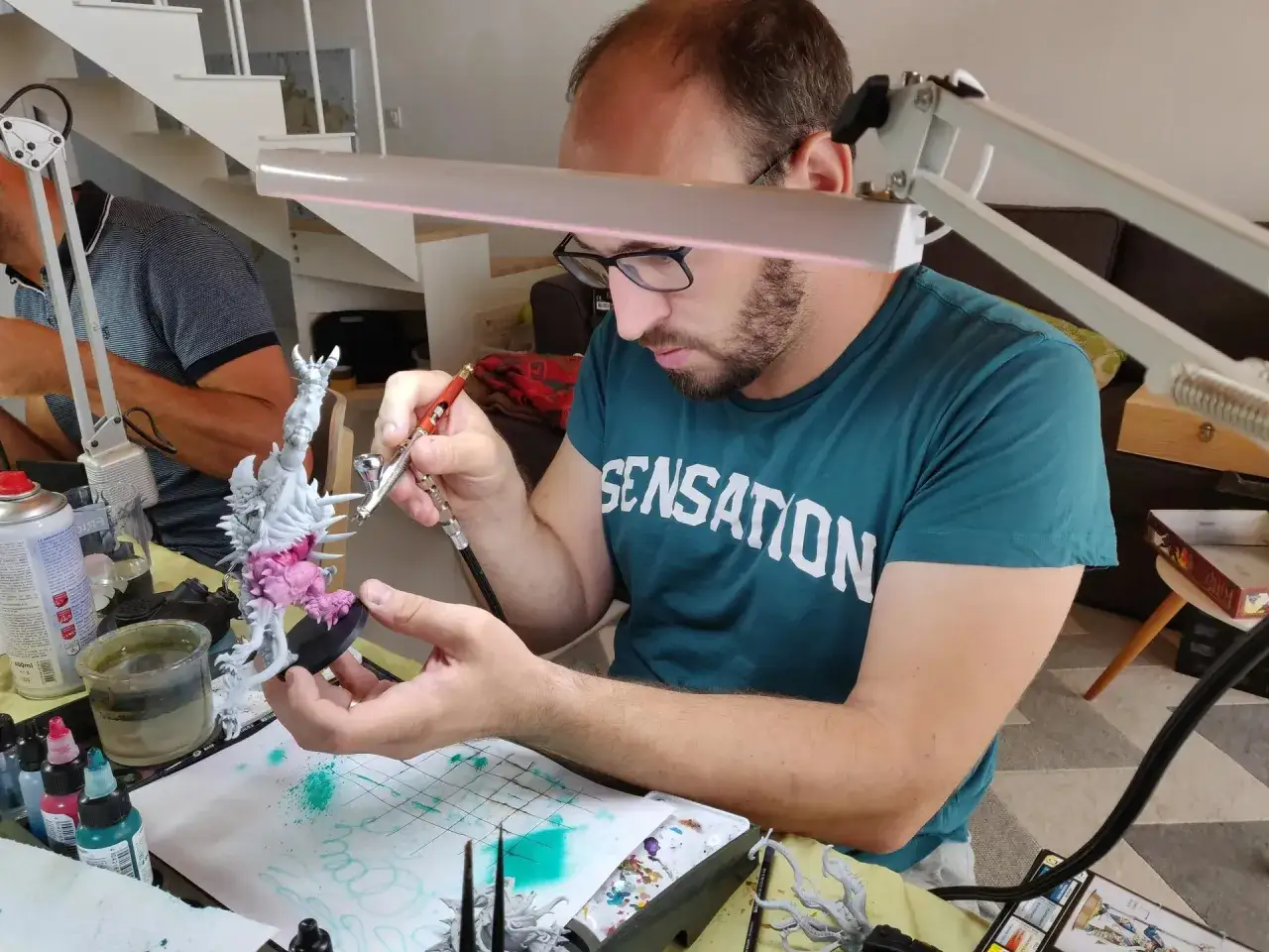 Un homme concentré apprend à peindre une figurine avec un aérographe. Des pots de peinture et des pinceaux sont disposés autour de lui.