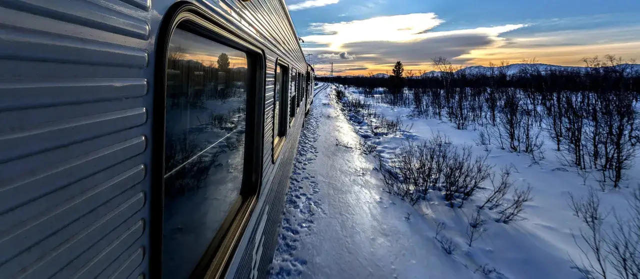Tåget rullar genom ett snöigt landskap mot Kiruna. Solnedgången färgar himlen och reflekteras i fönstren.