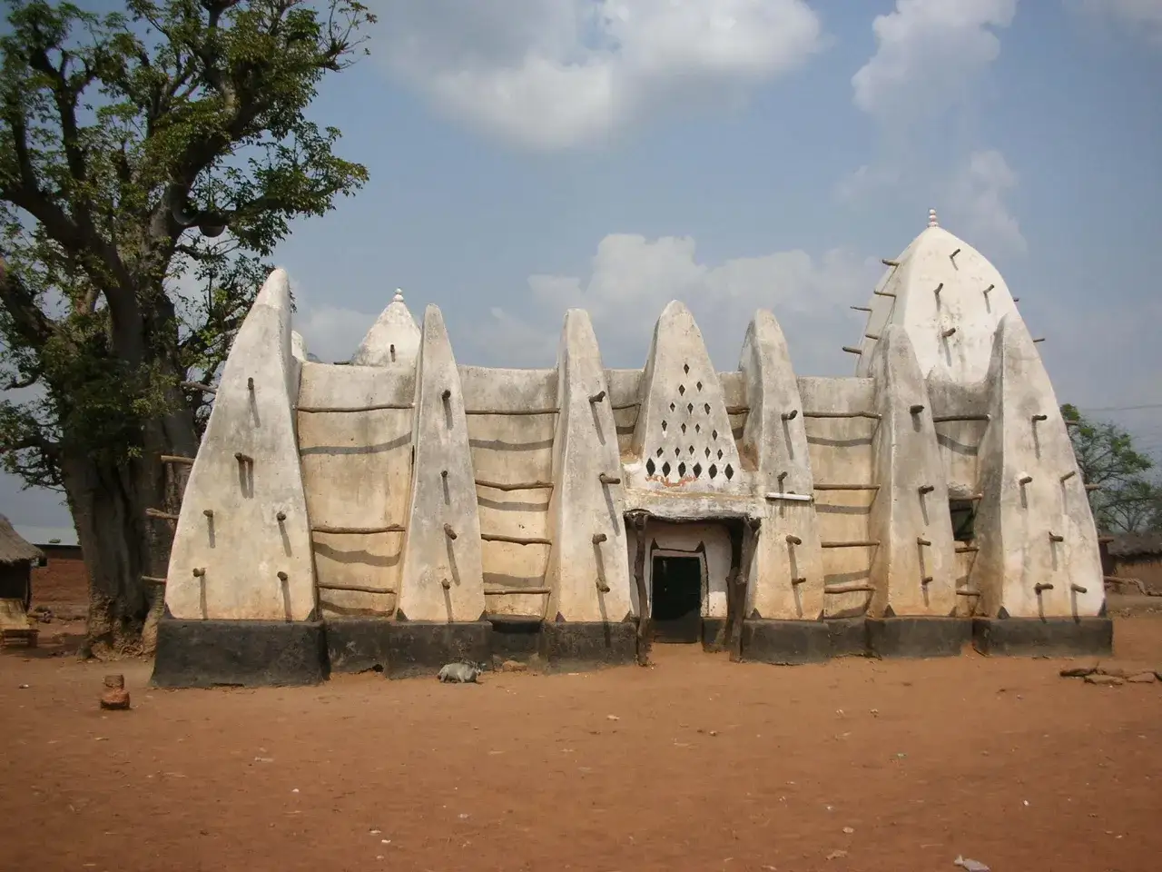 Tradycyjna budowla w **Ghana kraj**, z charakterystycznymi stożkowymi elementami i drewnianymi belkami, stoi obok potężnego drzewa baobab.