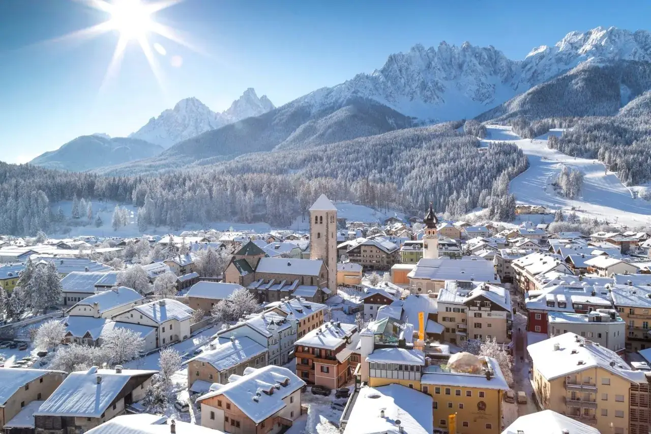 Un incantevole panorama di **Trento inverno**, con tetti innevati, montagne maestose e un sole splendente che illumina il borgo.