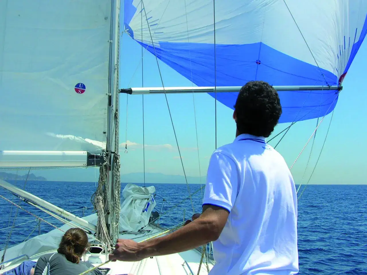 Uomo in barca a vela osserva il grande spinnaker blu e bianco gonfiarsi al vento, pronto per la regata.