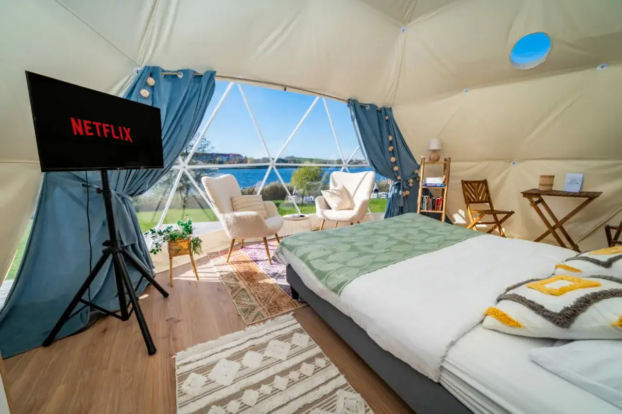 Wygodny camping wagabunda z Netflixem i widokiem na jezioro. Dwa fotele, łóżko i stolik tworzą przytulne wnętrze.
