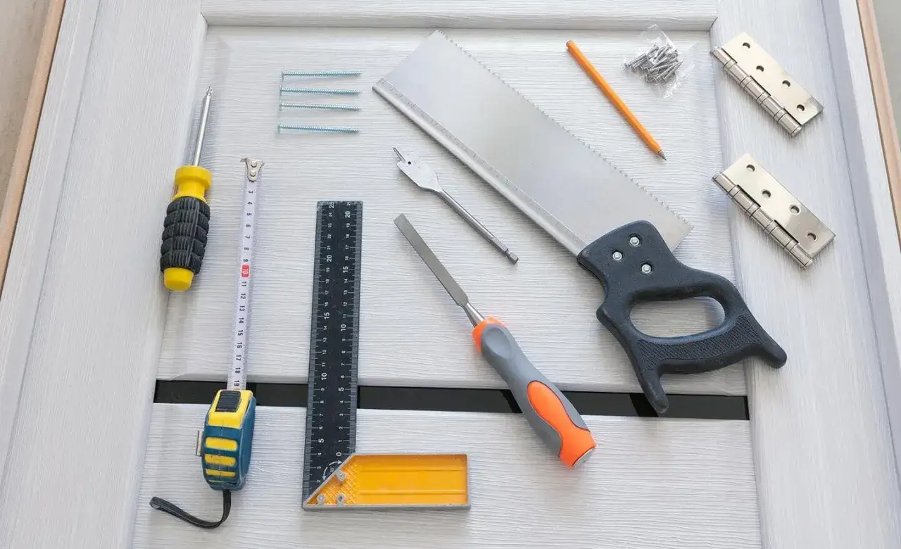 Tools laid out for how to hang a door: saw, screwdriver, tape measure, ruler, chisel, pencil, screws, and hinges on a white door.