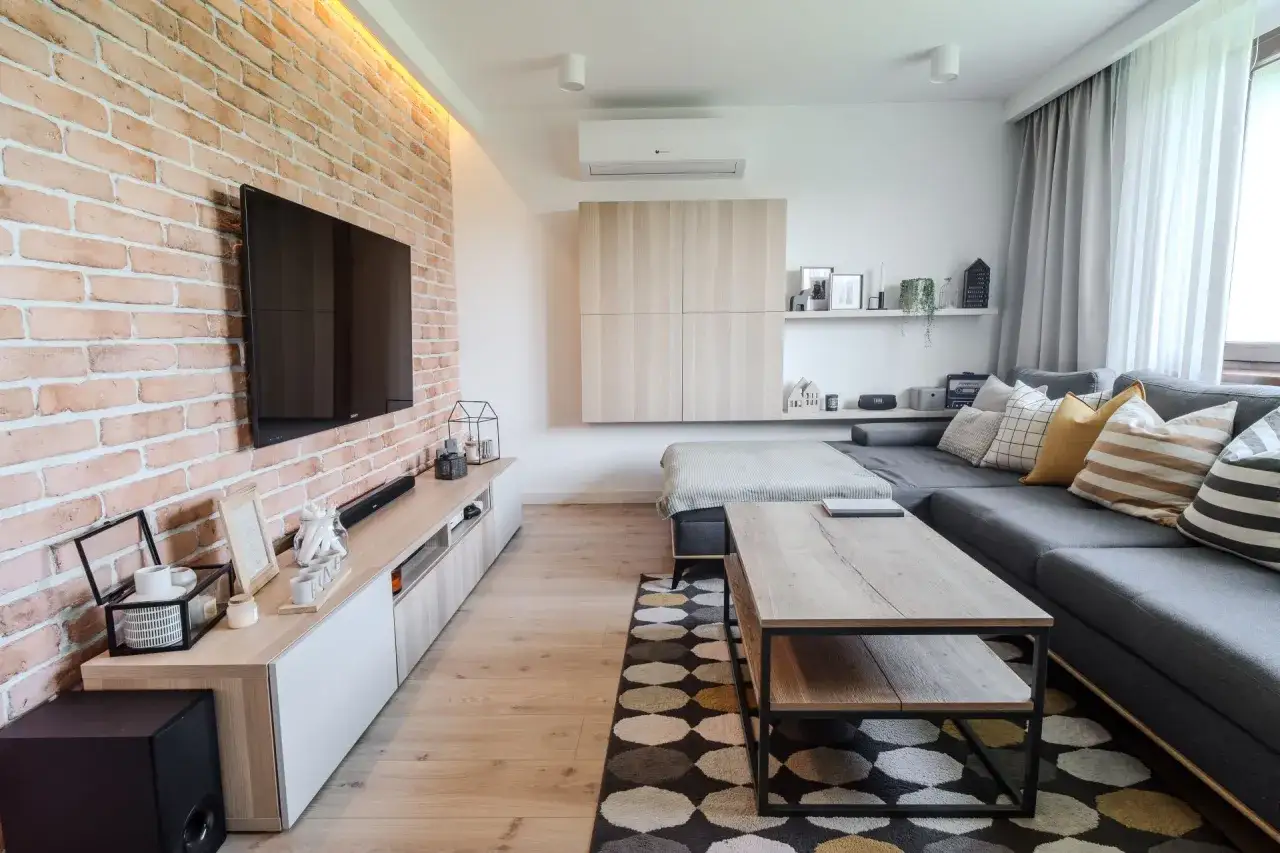 Nowoczesny salon w domu kostce. Metamorfoza wnętrza z ceglaną ścianą, szarą kanapą i drewnianymi meblami.