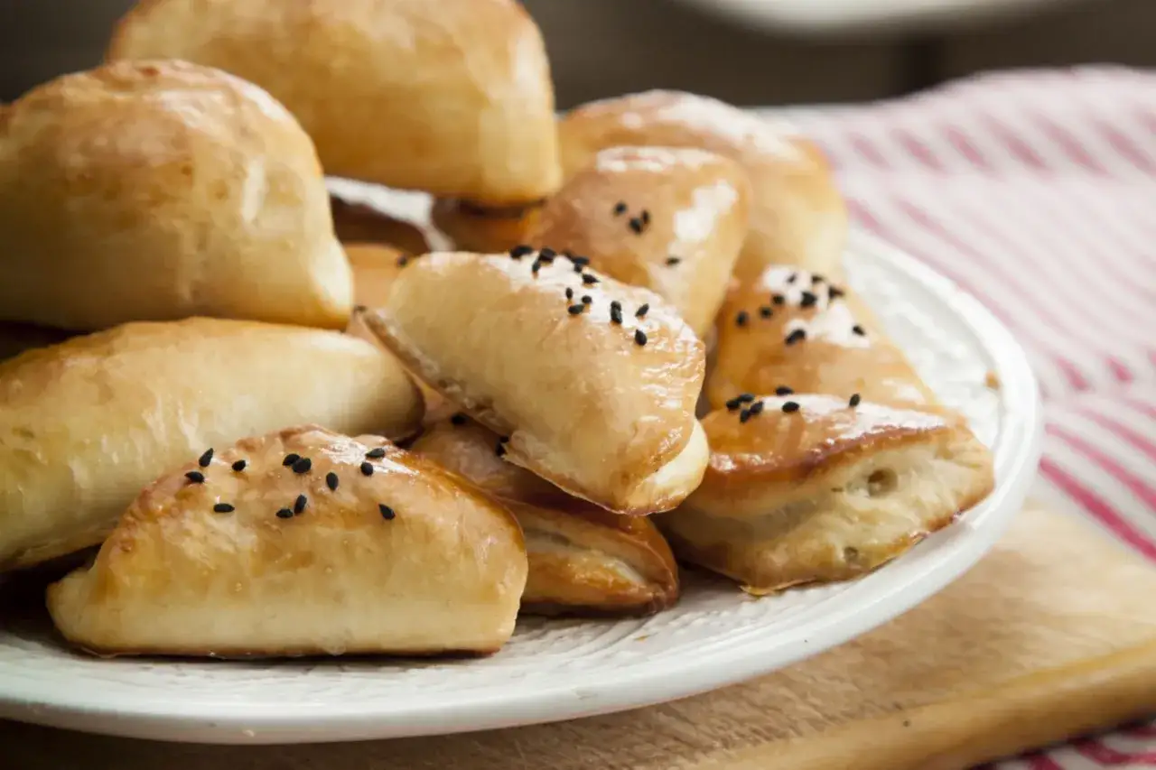Stos puszystych pierogów drożdżowych, niektóre posypane czarnuszką, na białym talerzu.