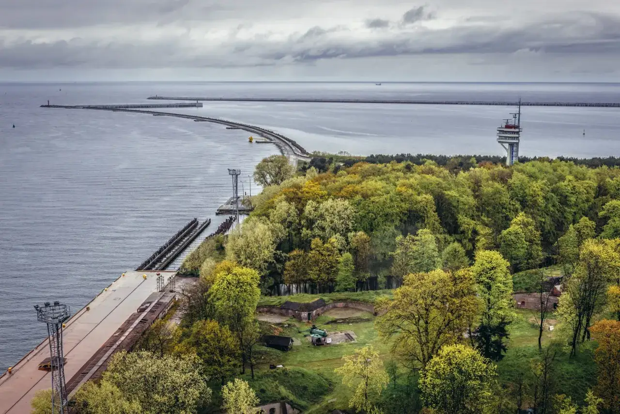 Widok na port i fortyfikacje w Świnoujściu. W oddali widać falochrony i wieżę pilot&oacute;w, a na pierwszym planie zielony las i ruiny fortu. To jedne z wielu świnoujskich atrakcji.