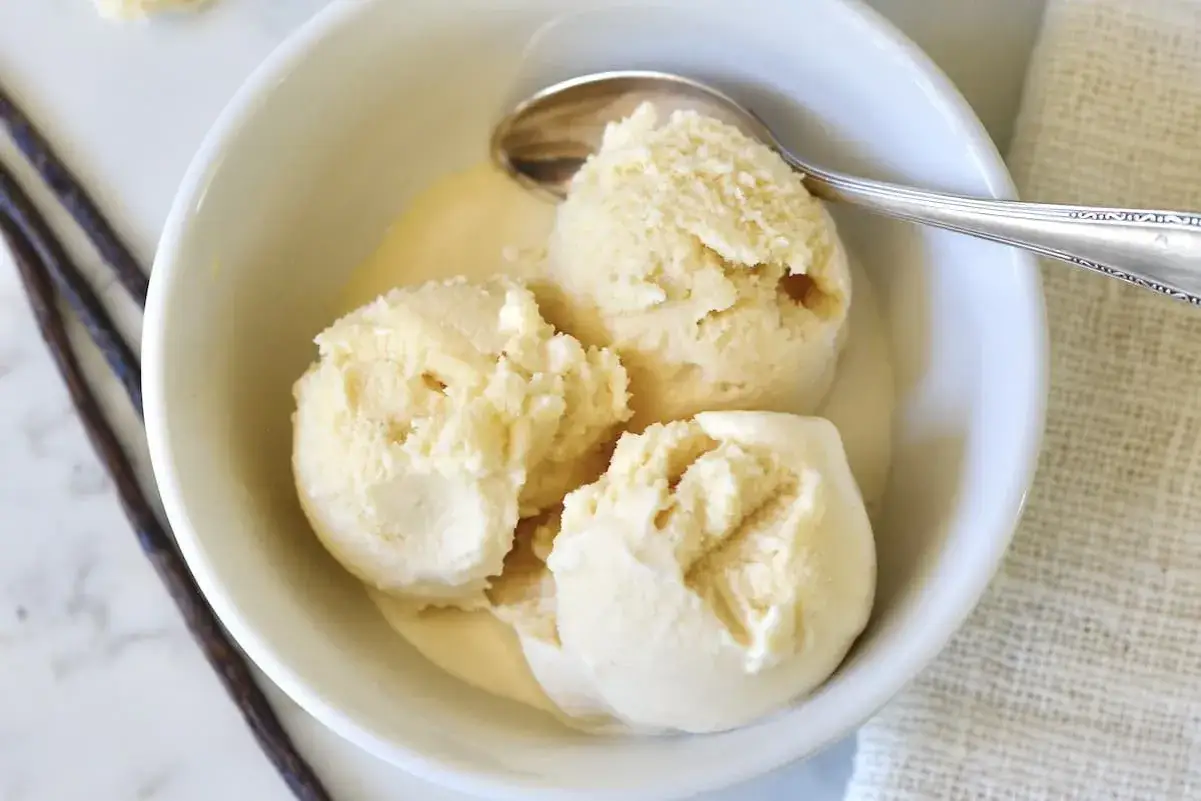 Cremiges Milch-Eis in einer Schüssel, geschmolzen und mit einem Löffel. Daneben liegen Vanilleschoten.