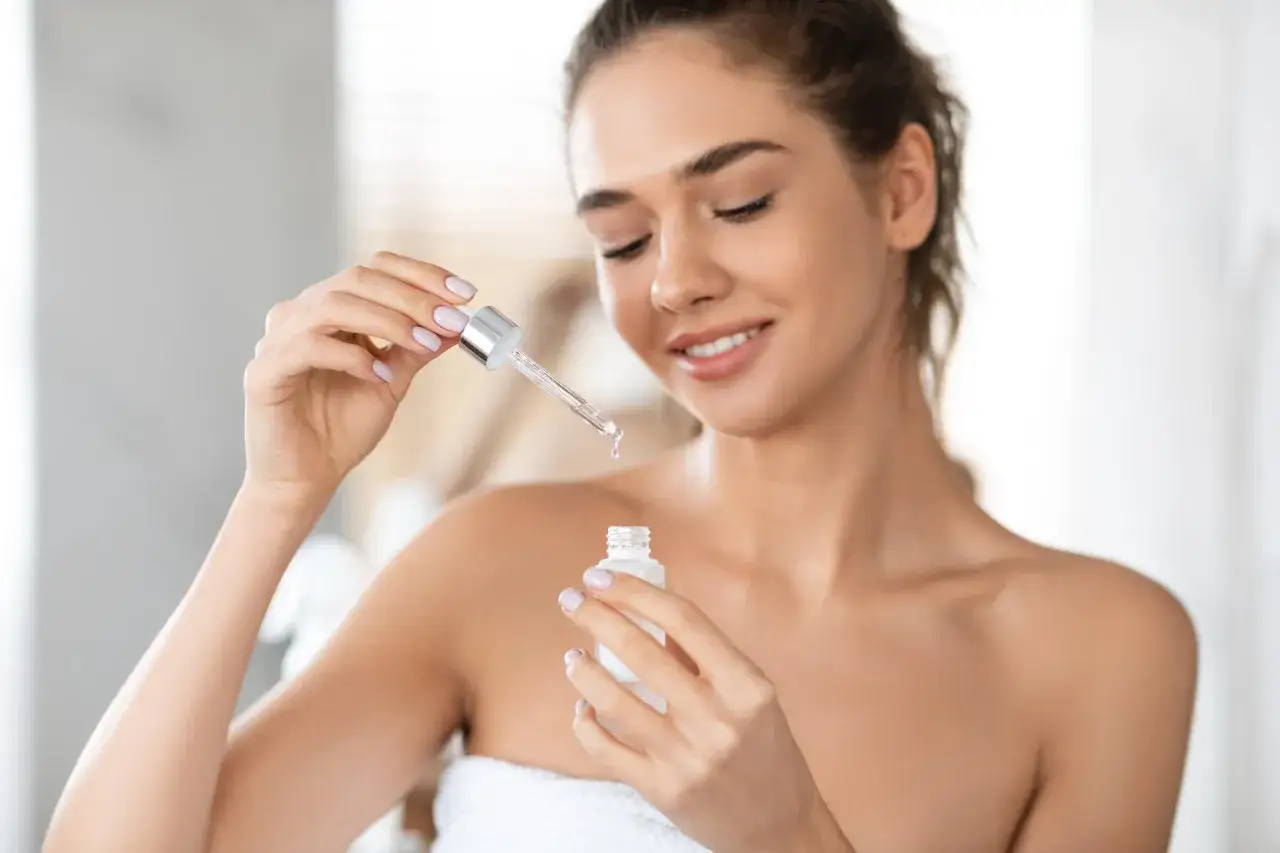 Kobieta z uśmiechem pokazuje, jak stosować serum do twarzy, trzymając w dłoniach pipetkę z kroplą produktu i małą buteleczkę.