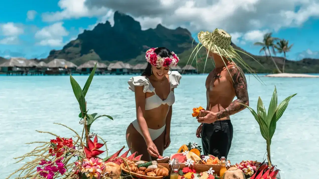 Ett par njuter av en frukost i vattnet p&aring; Franska Polynesien, med &ouml;vervattensbungalows och en bergig &ouml; i bakgrunden.
