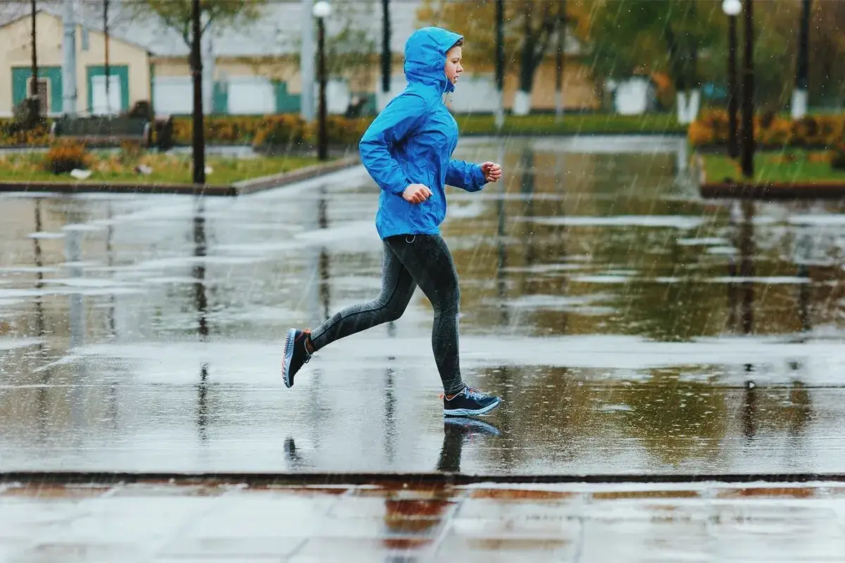 En person i bl&aring; jacka med huva springer i regnet p&aring; en bl&ouml;t trottoar.