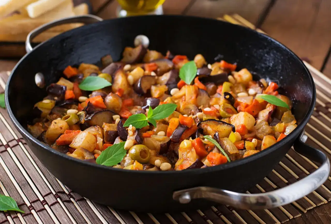 Caponata co to? Sycylijska potrawa z bakłażana, pomidorów, oliwek i orzeszków piniowych, podana w czarnej patelni.
