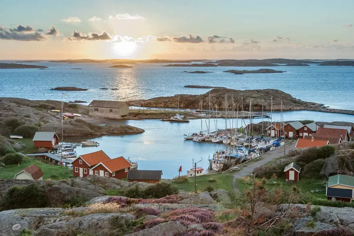 En svensk kustby med röda hus och segelbåtar i hamnen, badad i solnedgångens gyllene ljus. Perfekt för sommar vibes.