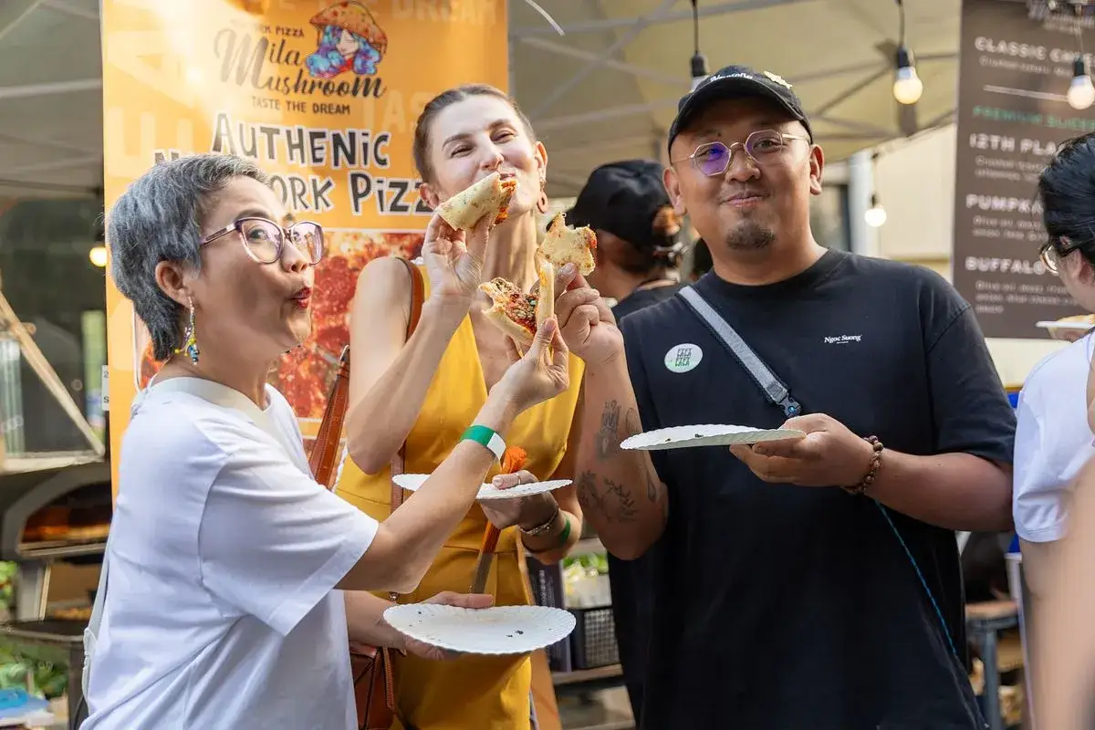 Ludzie cieszą się pizzą na festiwalu pizzy. Kobieta w żółtej sukience je kawałek, a mężczyzna w czarnej koszulce trzyma talerzyk z pizzą.