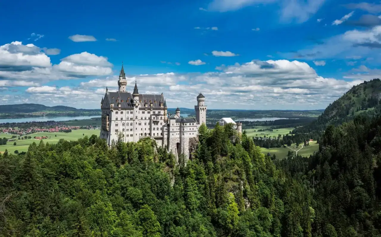 El majestuoso castillo de Neuschwanstein se alza sobre un acantilado verde, con torres que tocan un cielo azul y nubes blancas.