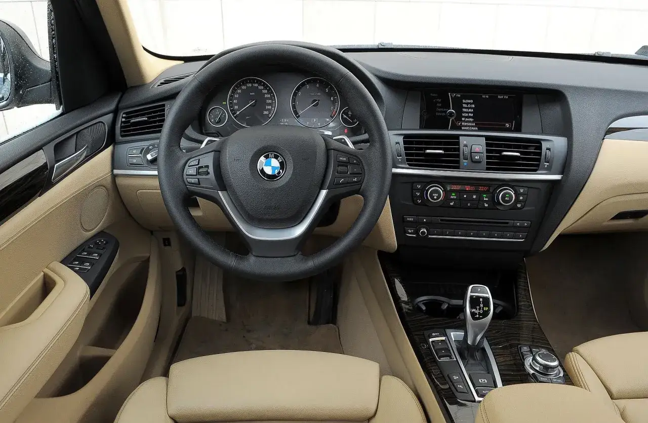 Wnętrze BMW X3 E83 z beżową tapicerką, kierownicą wielofunkcyjną i panelem radia.