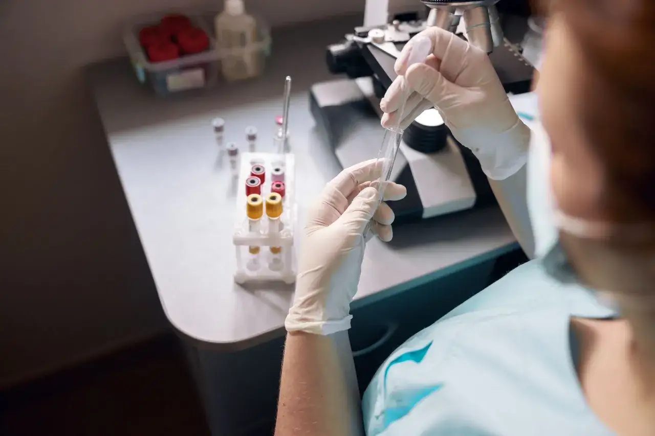 W tym laboratorium naukowiec w rękawiczkach pobiera próbkę pipetą do probówki, obok stoją inne probówki i mikroskop.