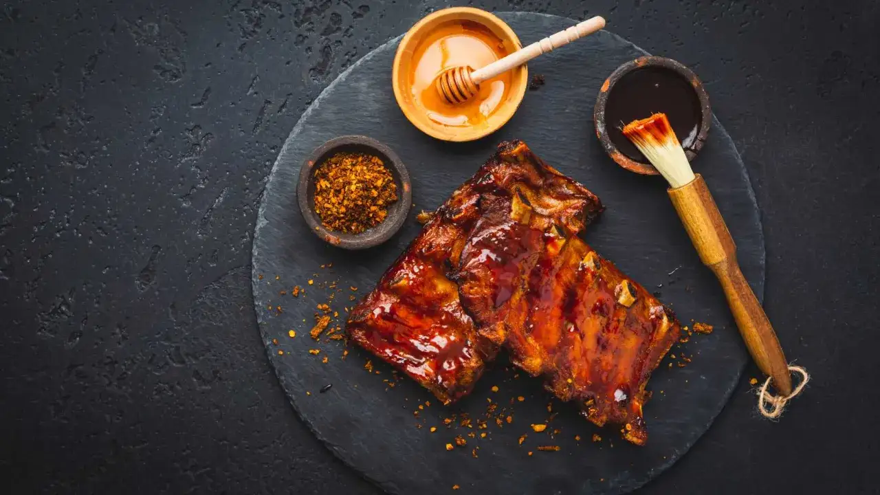 Costillas al horno glaseadas con miel y salsa barbacoa, acompañadas de especias y un pincel para untar.