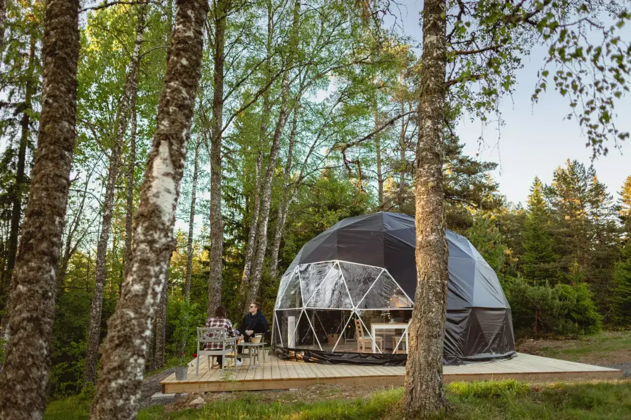 Tv&aring; personer sitter vid ett bord utanf&ouml;r en glampingkupol i skogen. En mysig plats att campa i skogen.