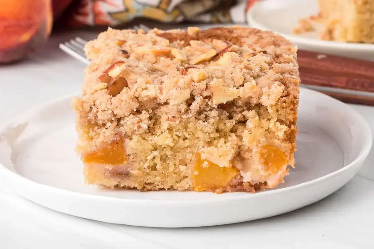 Ein Stück saftiger Pfirsichkuchen mit Streuseln und Mandeln auf einem weißen Teller.