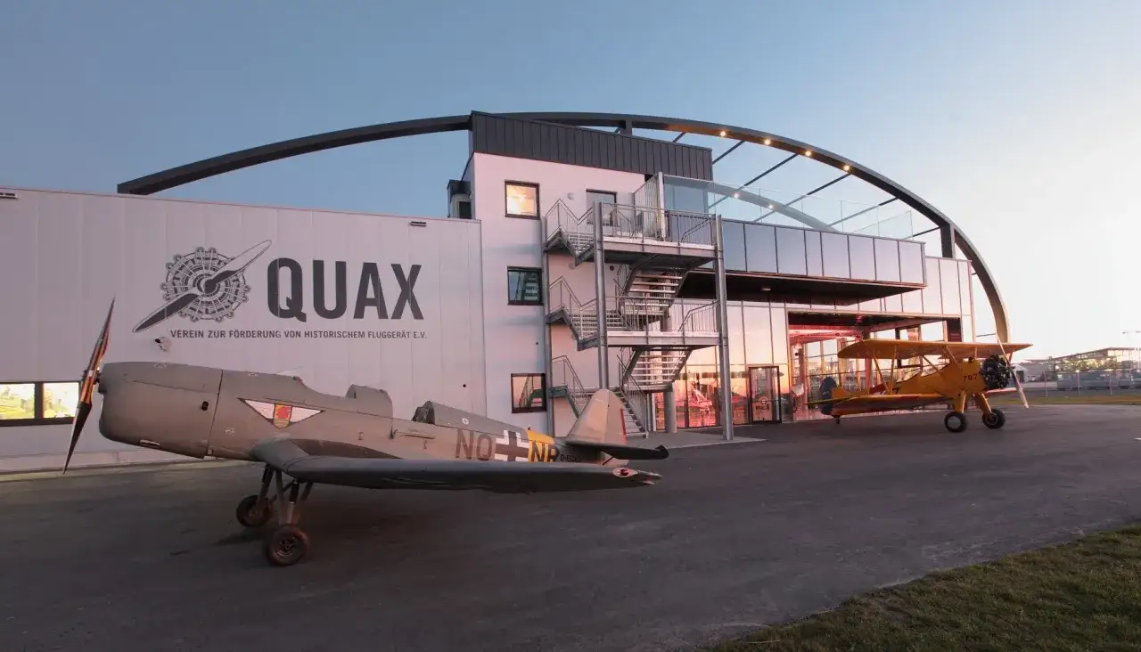 Zwei historische Flugzeuge stehen vor dem Quax Hangar. Ein graues Flugzeug mit Propeller im Vordergrund, ein gelbes Doppeldeckerflugzeug daneben.