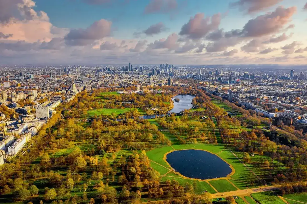 Jesień w Hyde Park w Londynie. Złote liście drzew otaczają stawy, a w tle widać panoramę miasta.