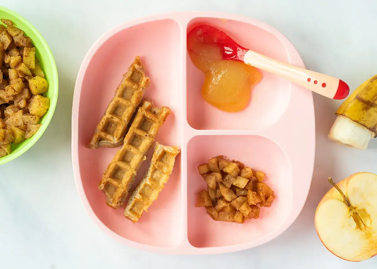 Waffelteig mit Apfelmus auf einem Kindergeschirr. Daneben Apfelstücke und eine Banane.