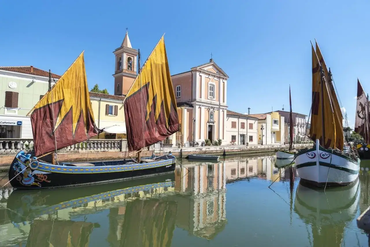 Barche tradizionali con vele colorate ormeggiate sul canale, sullo sfondo la chiesa e le case della riviera Cesenatico.