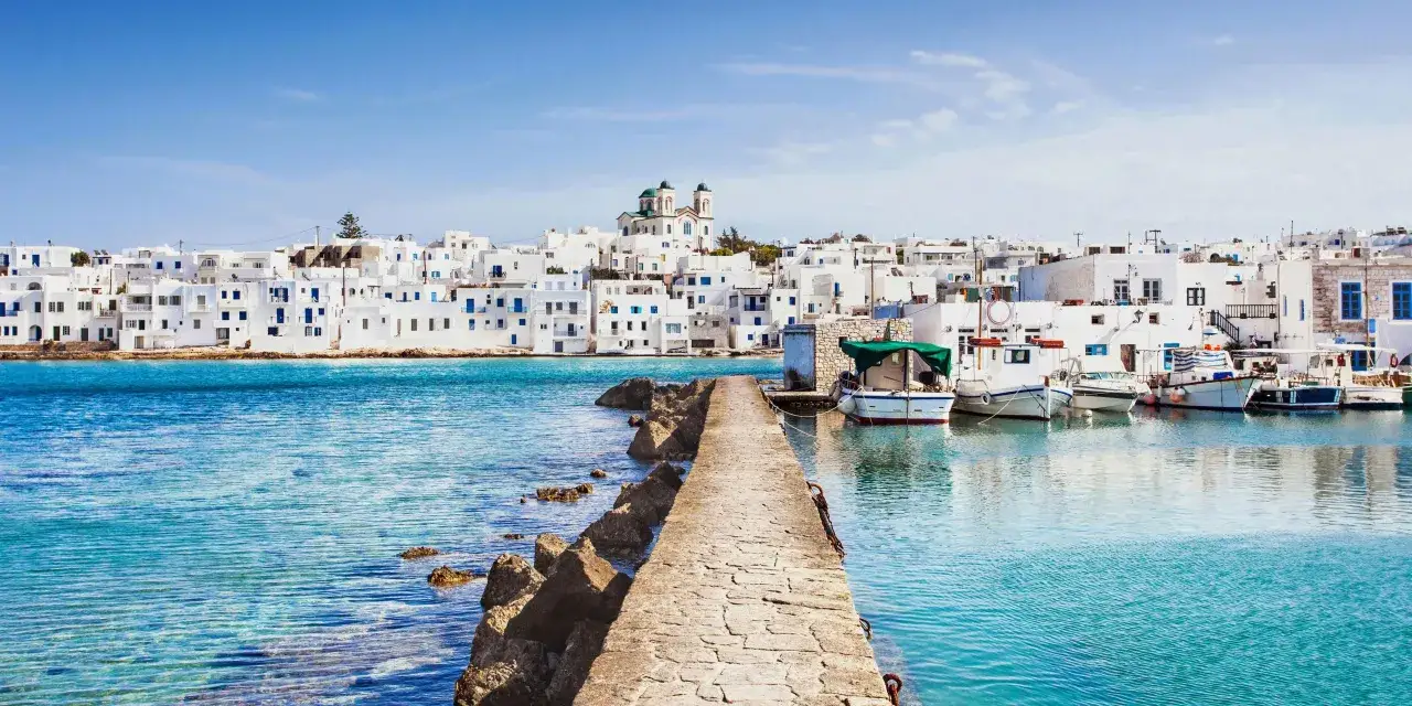 Ein Pier f&uuml;hrt zu einem malerischen Hafen auf der Insel Paros, Griechenland. Wei&szlig;e H&auml;user mit blauen Akzenten s&auml;umen die K&uuml;ste, Boote schaukeln im t&uuml;rkisfarbenen Wasser.