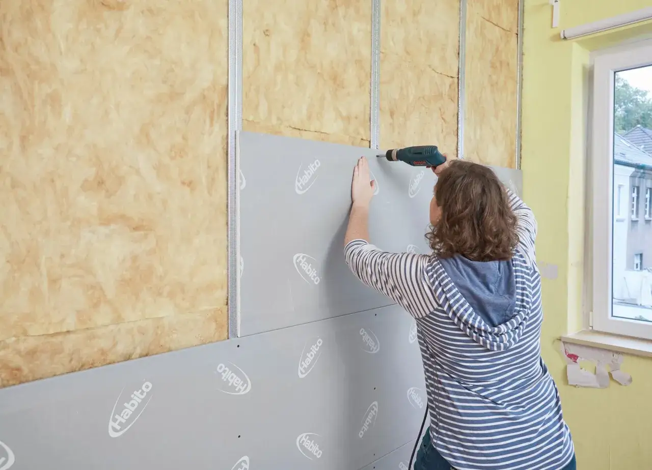 Frau schraubt Rigipsplatten an eine Wand mit Dämmung. Die Platten werden mit einem Akkuschrauber befestigt.