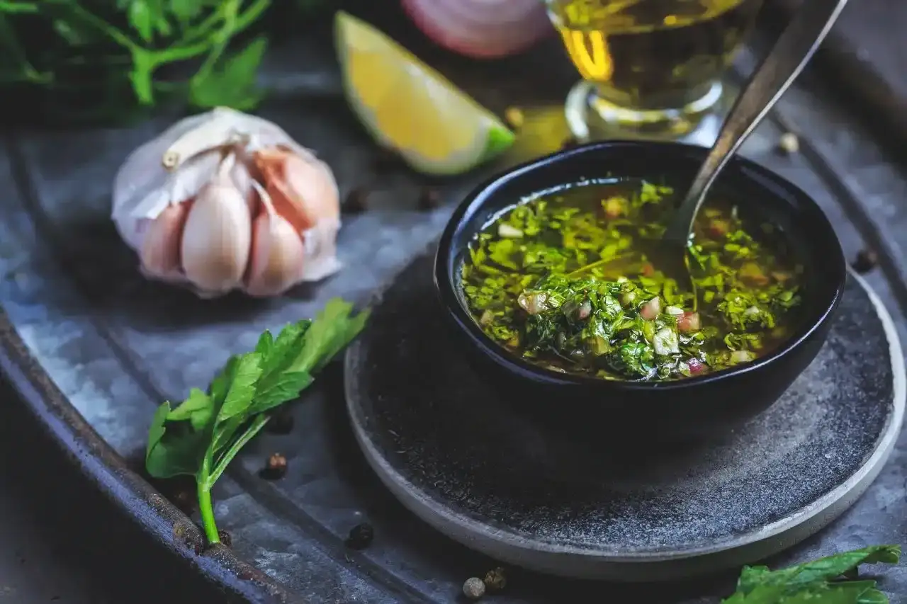 Aromatyczne chimichuri w miseczce, obok czosnek, limonka i oliwa. Idealny dodatek do grillowanych mięs.