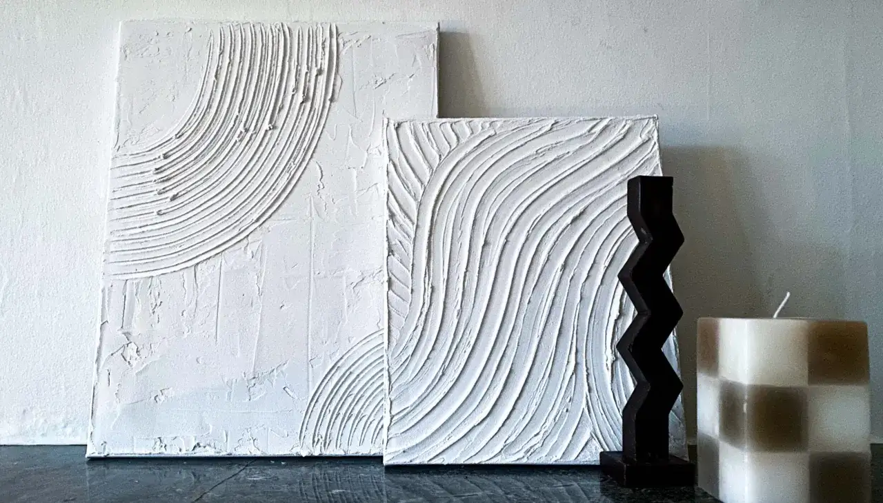 Två vita texturtavlor, skapade för att måla tavla med spackel, står bredvid en mörk skulptur och en randig ljus.