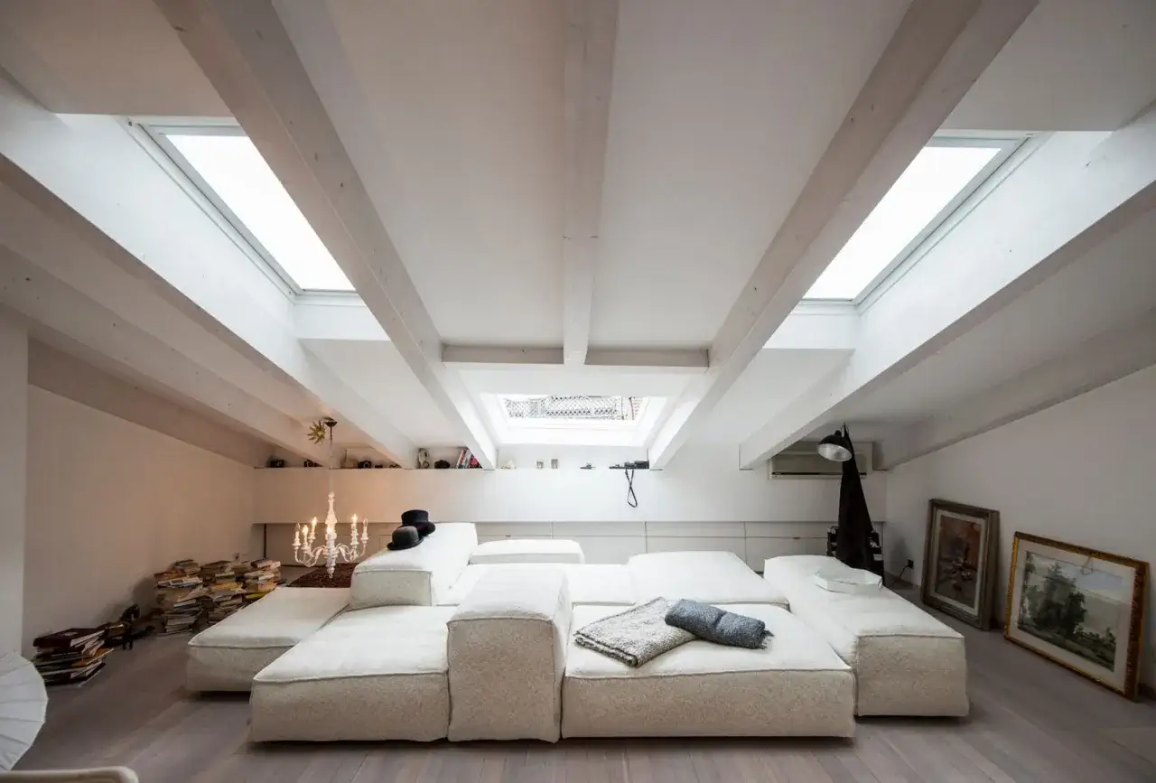 Soffitto con travi in legno di bianco, lucernari e un ampio divano modulare bianco.
