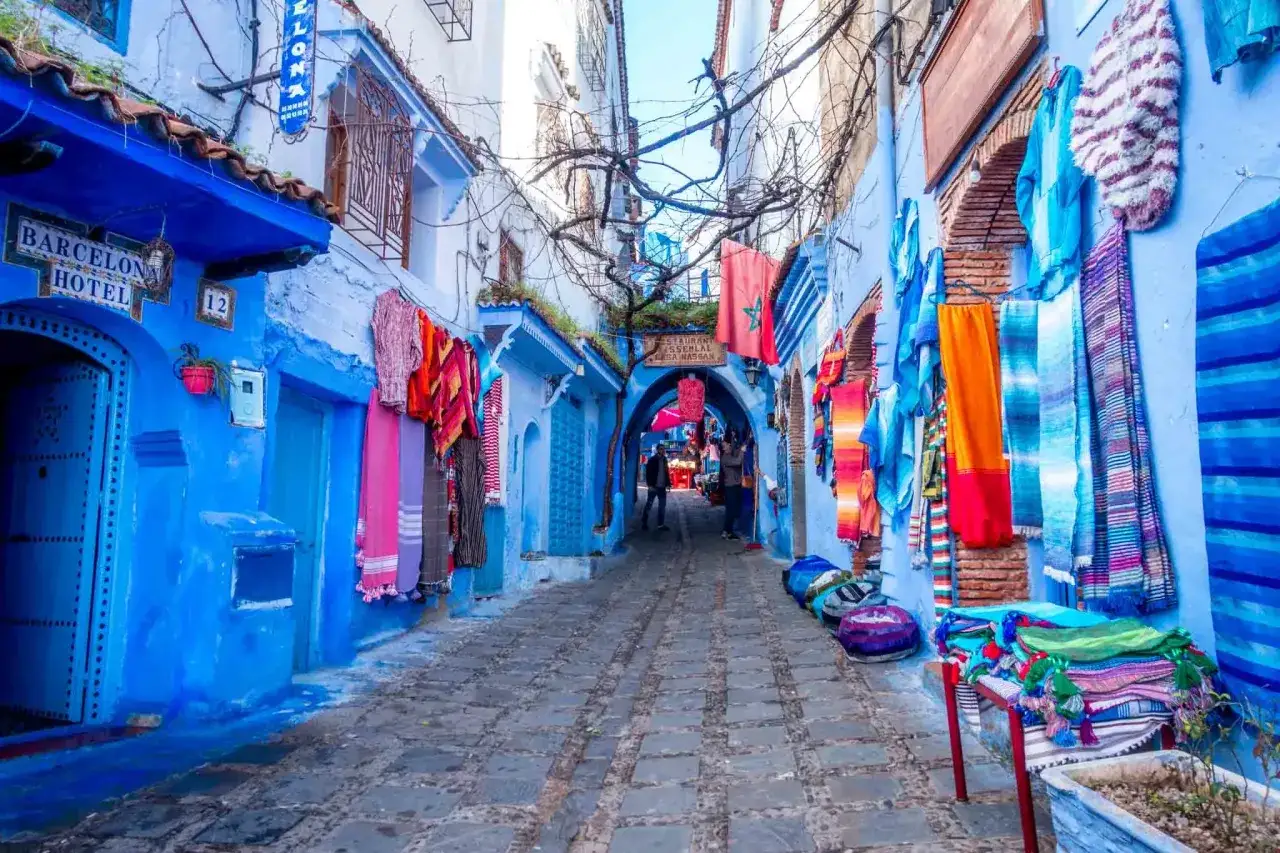 Blaue Gassen in Chefchaouen, warum blau? Bunte Textilien h&auml;ngen an den W&auml;nden, ein Hotel-Schild ist zu sehen.