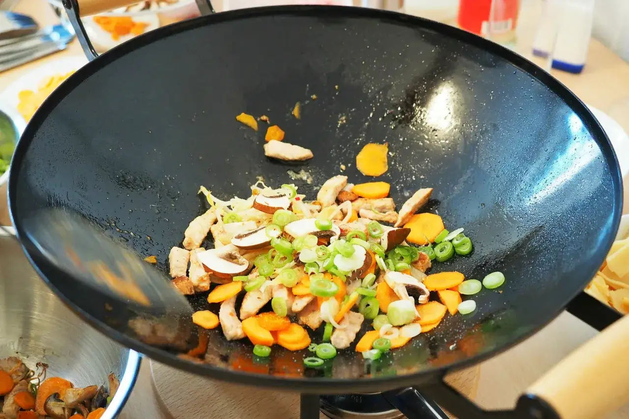 Na patelni wok smażą się kawałki kurczaka, marchewka, pieczarki i szczypiorek. Idealne składniki do szybkich wok przepisów.