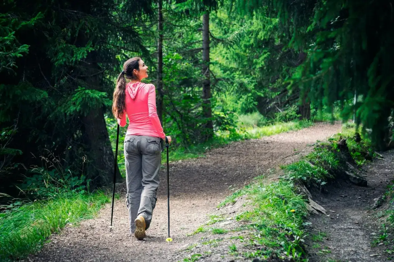 Kvinna går med stavar på en grusväg i en grönskande skog.