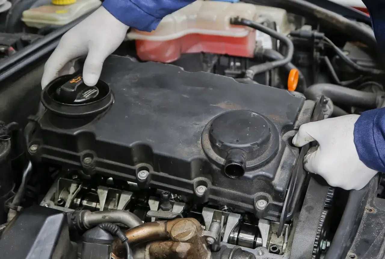 Mechanik w białych rękawiczkach sprawdza silnik, odkręcając korek wlewu oleju. Widoczny jest element odmy olejowej.