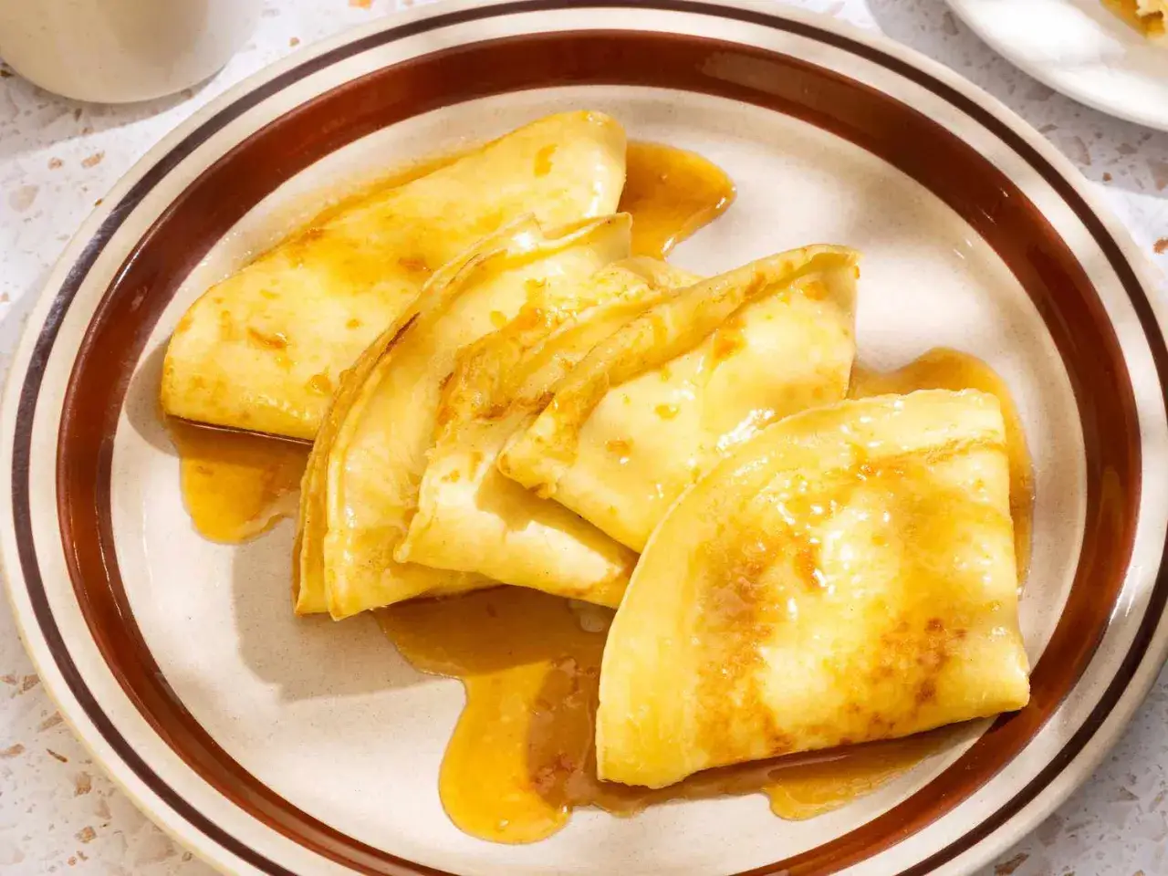 Złociste crepes suzette polane syropem, podane na talerzu z brązowym rantem.