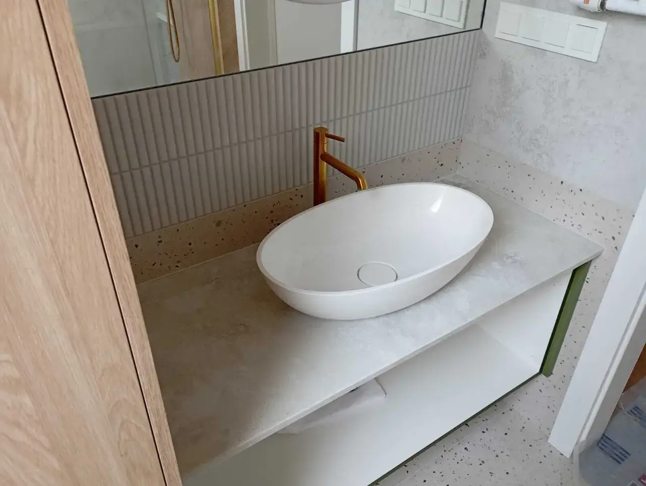 Elegancka łazienka z owalną umywalką nablatową i złotą baterią. Blat wykonany z jasnego konglomeratu kwarcowego z subtelnym wzorem.
