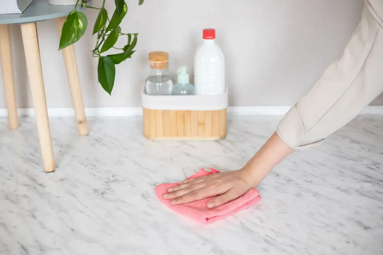A hand wipes a marble floor tile with a pink cloth, showing how to clean marble floor tiles. Cleaning supplies are in the background.